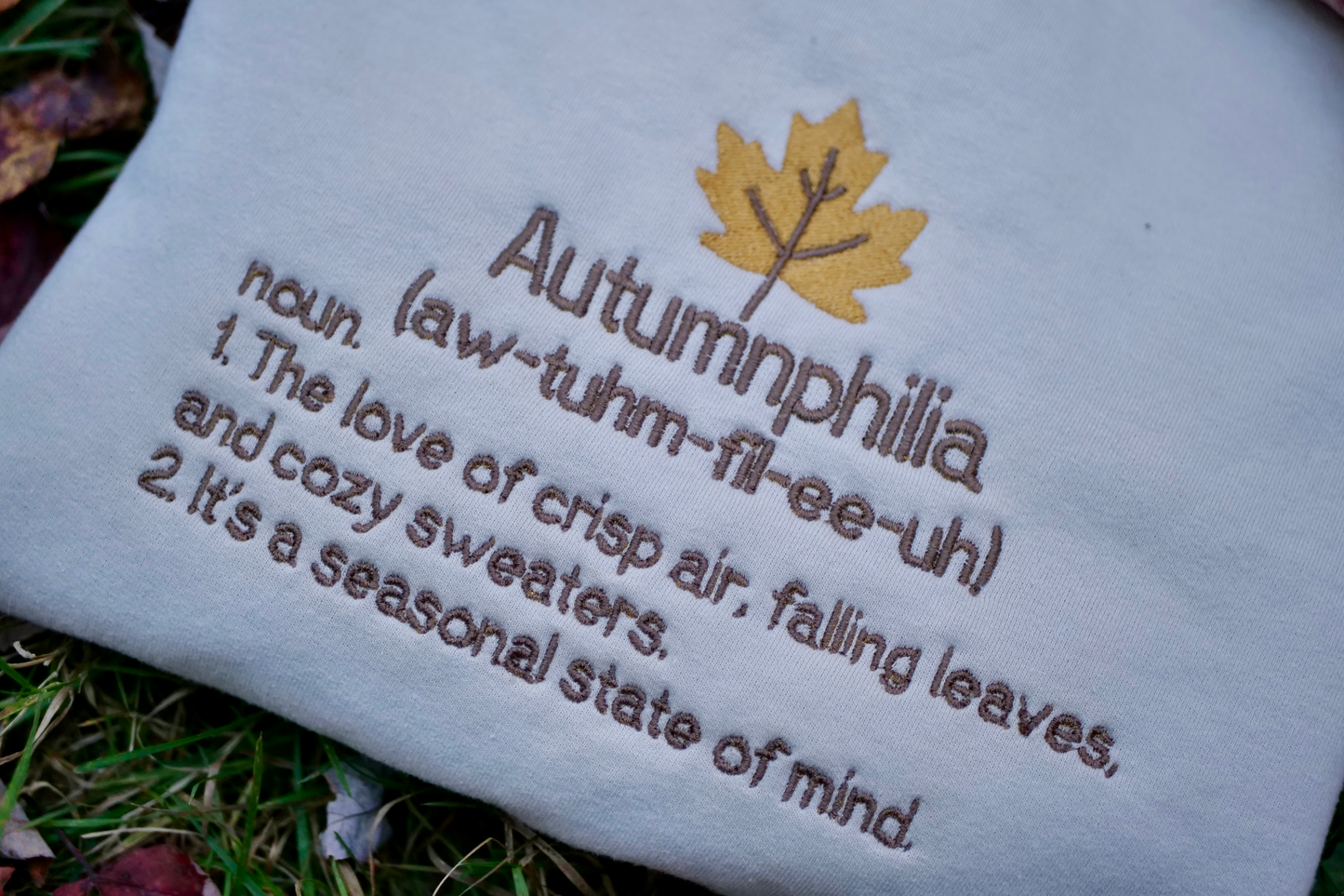 Textile with embroidered definition of 'Autumnphilia' and a maple leaf design on grass.