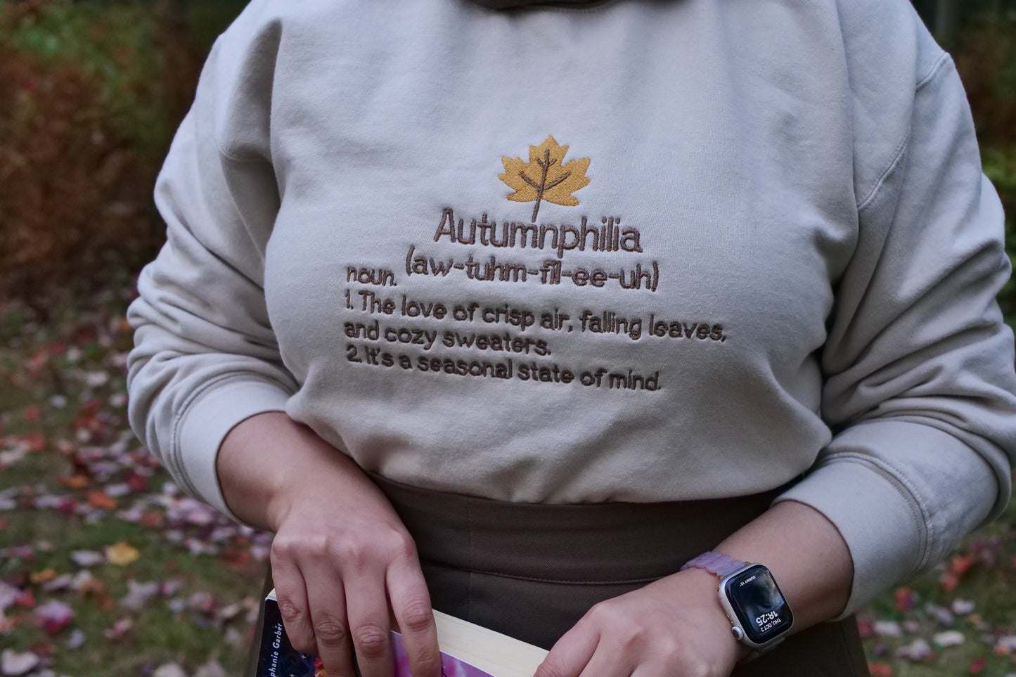 Person wearing a sweatshirt with 'Autumnphilia' definition and maple leaf design outdoors.