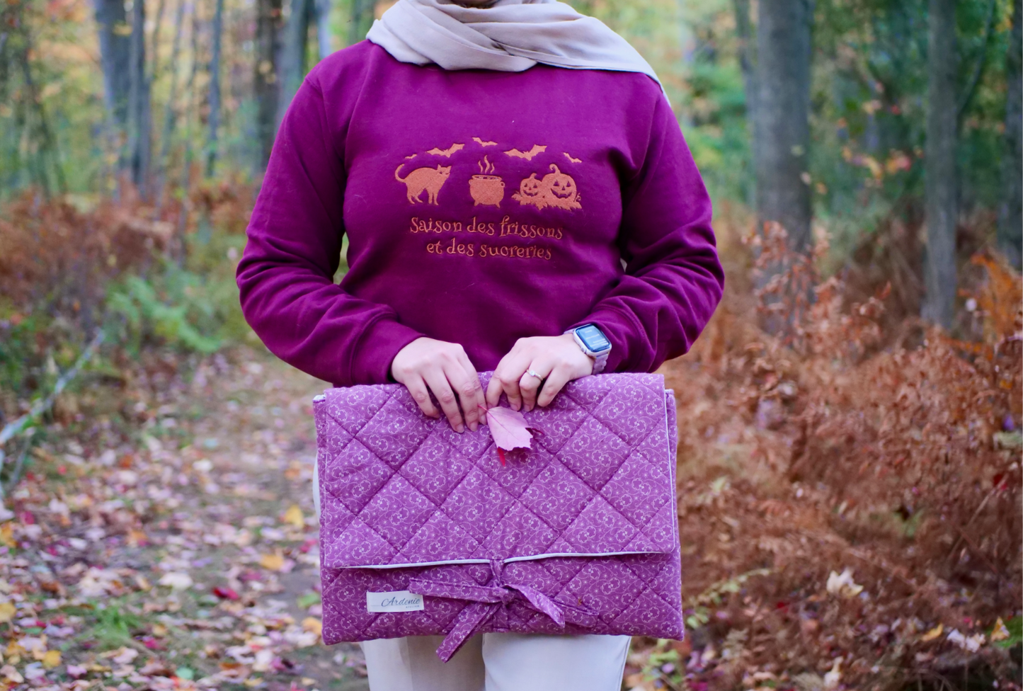 Person wearing a purple sweatshirt holding a matching purple quilted bag in a forest setting