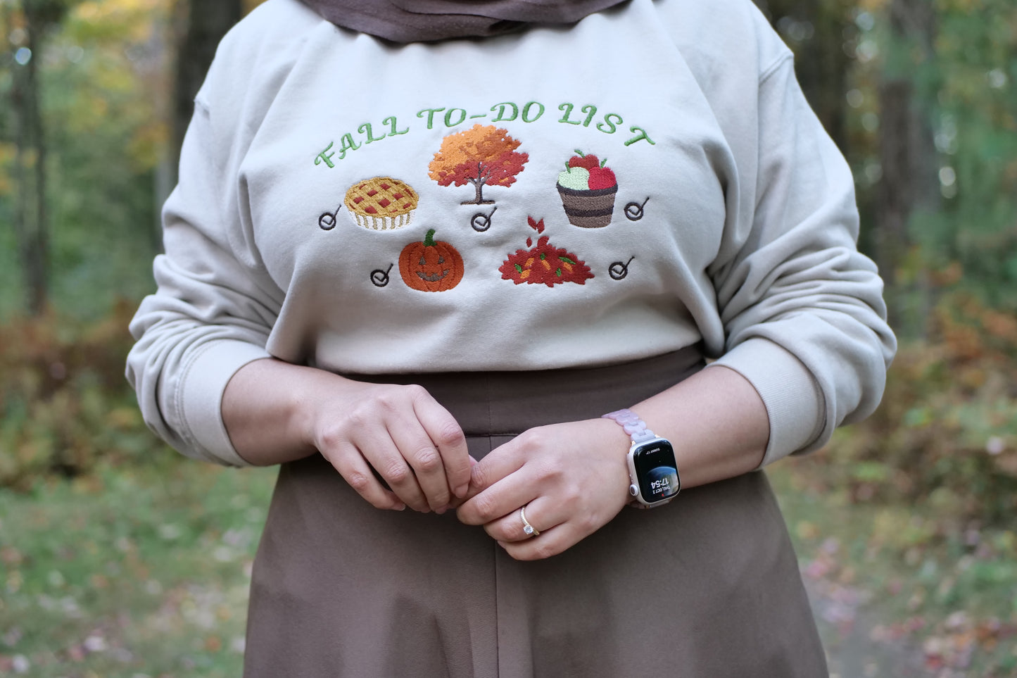 Person wearing a beige sweatshirt with 'Fall to Do List' text and autumn-themed graphics, standing in a forest.