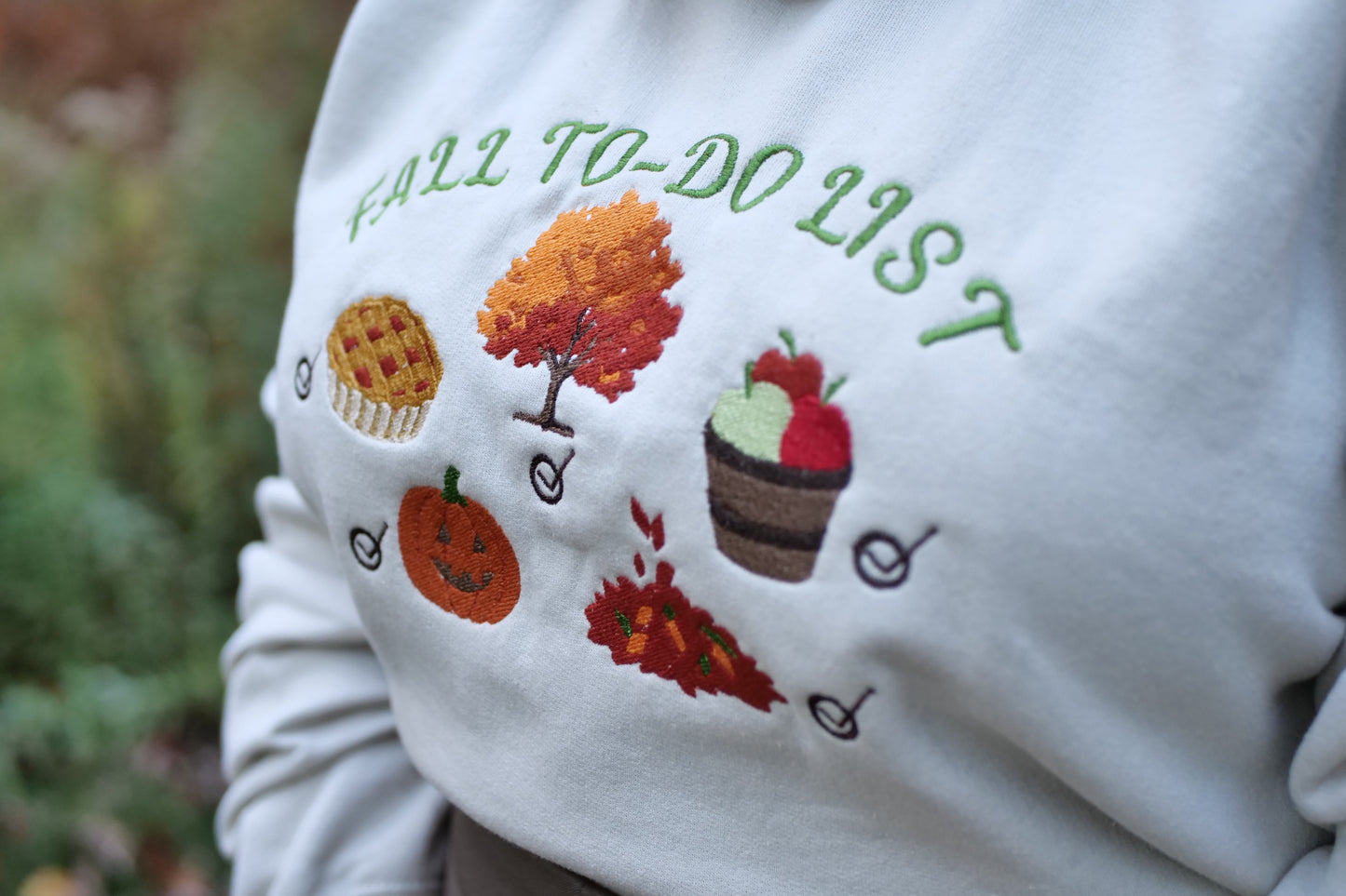 sand sweatshirt with autumn-themed embroidery and text 'FALL TO-DO LIST' on a blurred natural background.