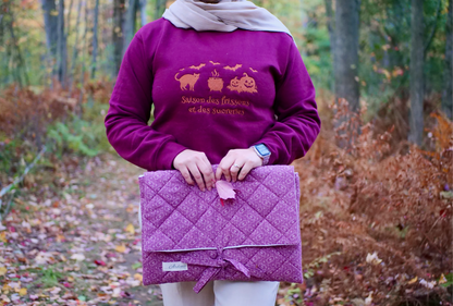 Person wearing a purple sweatshirt holding a matching purple quilted bag in a forest setting