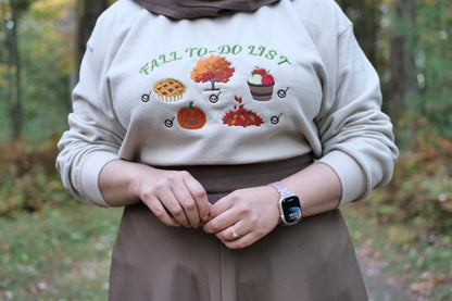 Person wearing a beige sweatshirt with 'Fall to Do List' text and autumn-themed graphics, standing in a forest.